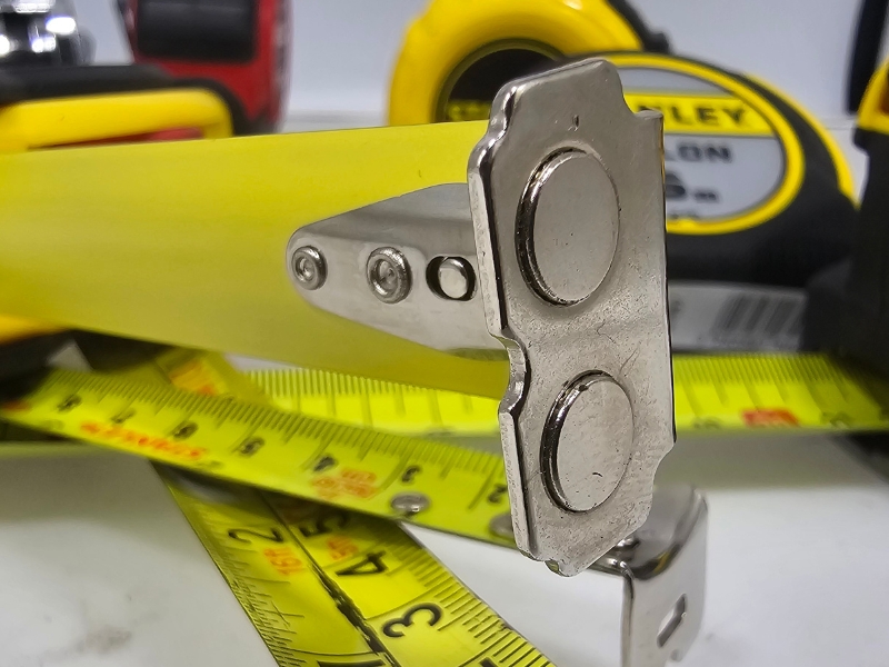 Zbliżenie na magnetyczny zaczep stalowej miarki zwijanej Duże zbliżenie stalowego zaczepu z wbudowanym magnesem na końcu taśmy miarki zwijanej, pokazujące szczegóły konstrukcji
